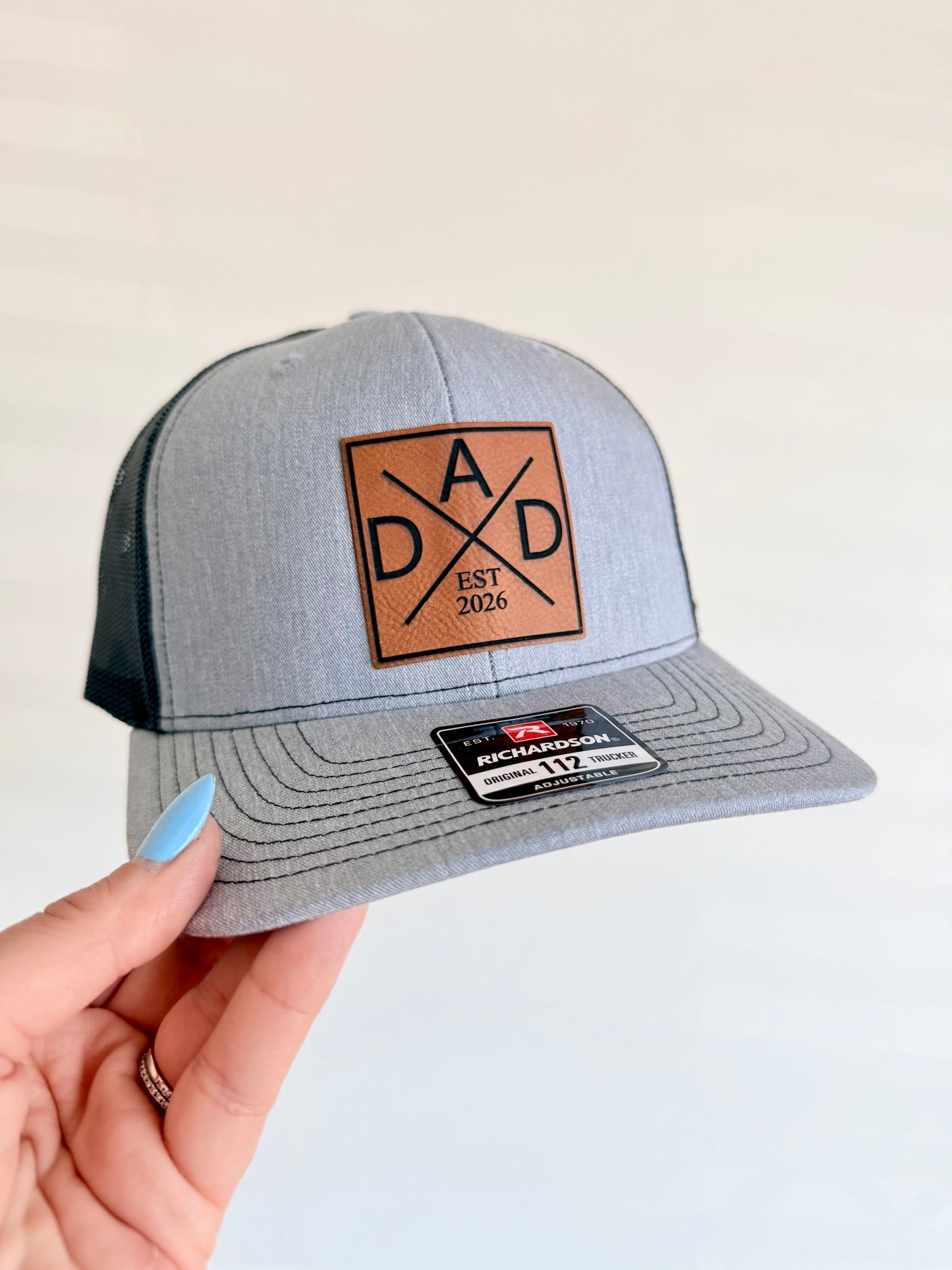 Gray cap with leather patch held by a hand on a light background. DAD Est. Custom laser engraved patch hat.