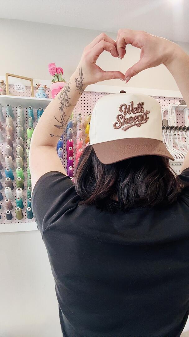 Person wearing a cap with 'Well Sheeple' text, making a heart shape with their hands in front of a wall of colorful bottles.