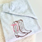 A folded light heather gray sweatshirt featuring an embroidered design of brown western cowboy boots with a pink bow on the side, displayed on a soft beige textured surface.