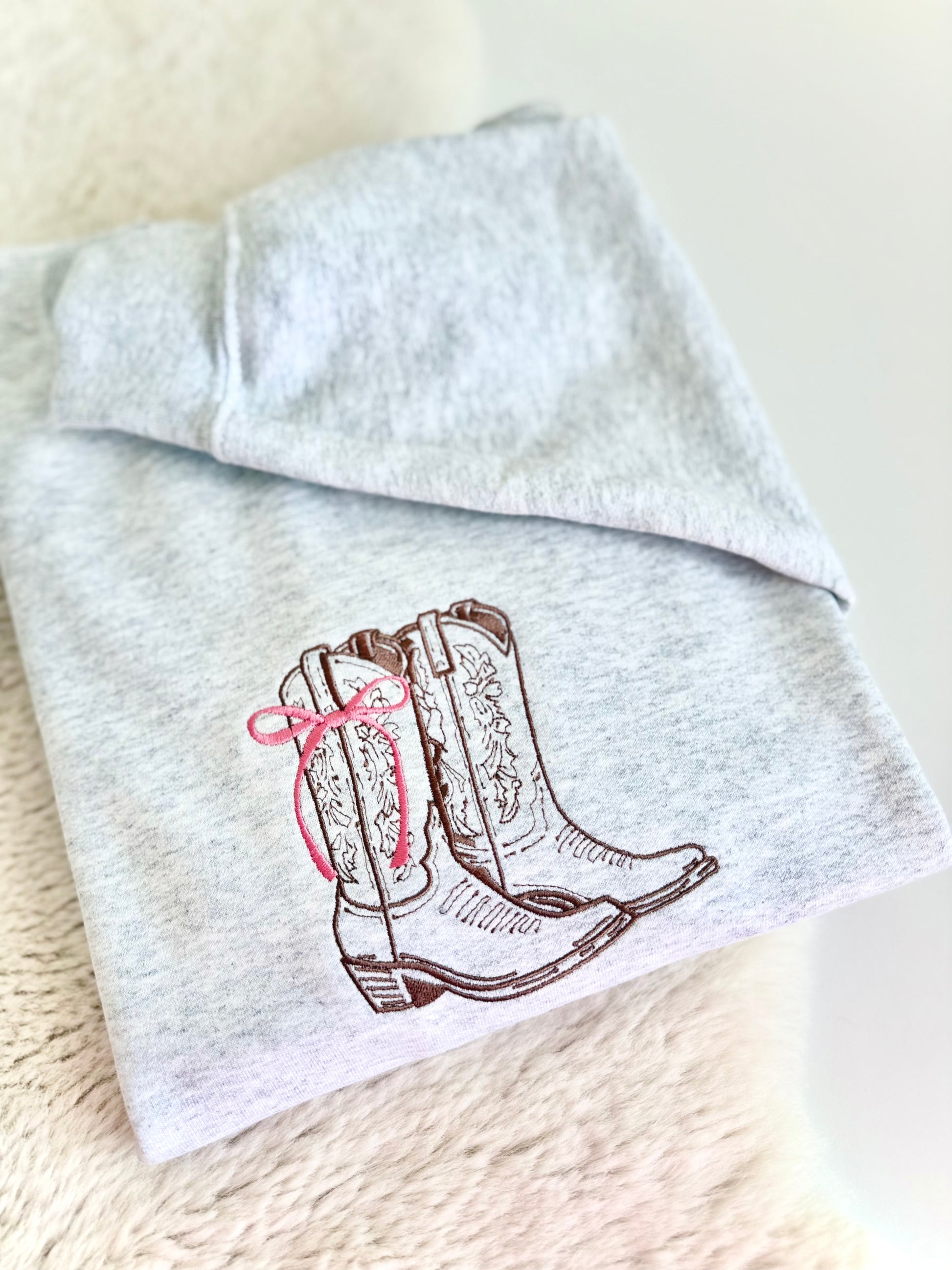 A folded light heather gray sweatshirt featuring an embroidered design of brown western cowboy boots with a pink bow on the side, displayed on a soft beige textured surface.