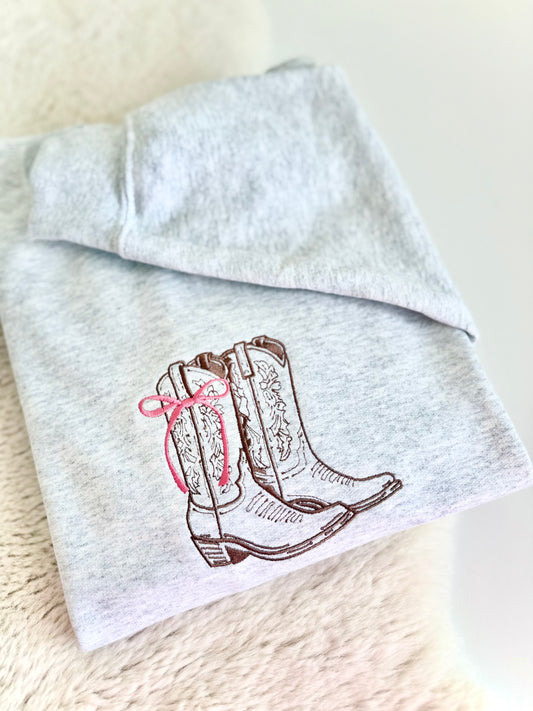 A folded light heather gray sweatshirt featuring an embroidered design of brown western cowboy boots with a pink bow on the side, displayed on a soft beige textured surface.