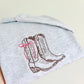 A folded light heather gray sweatshirt or hoodie with an embroidered design of cowboy boots in brown and a pink bow accent, held by a hand against a plain light background.