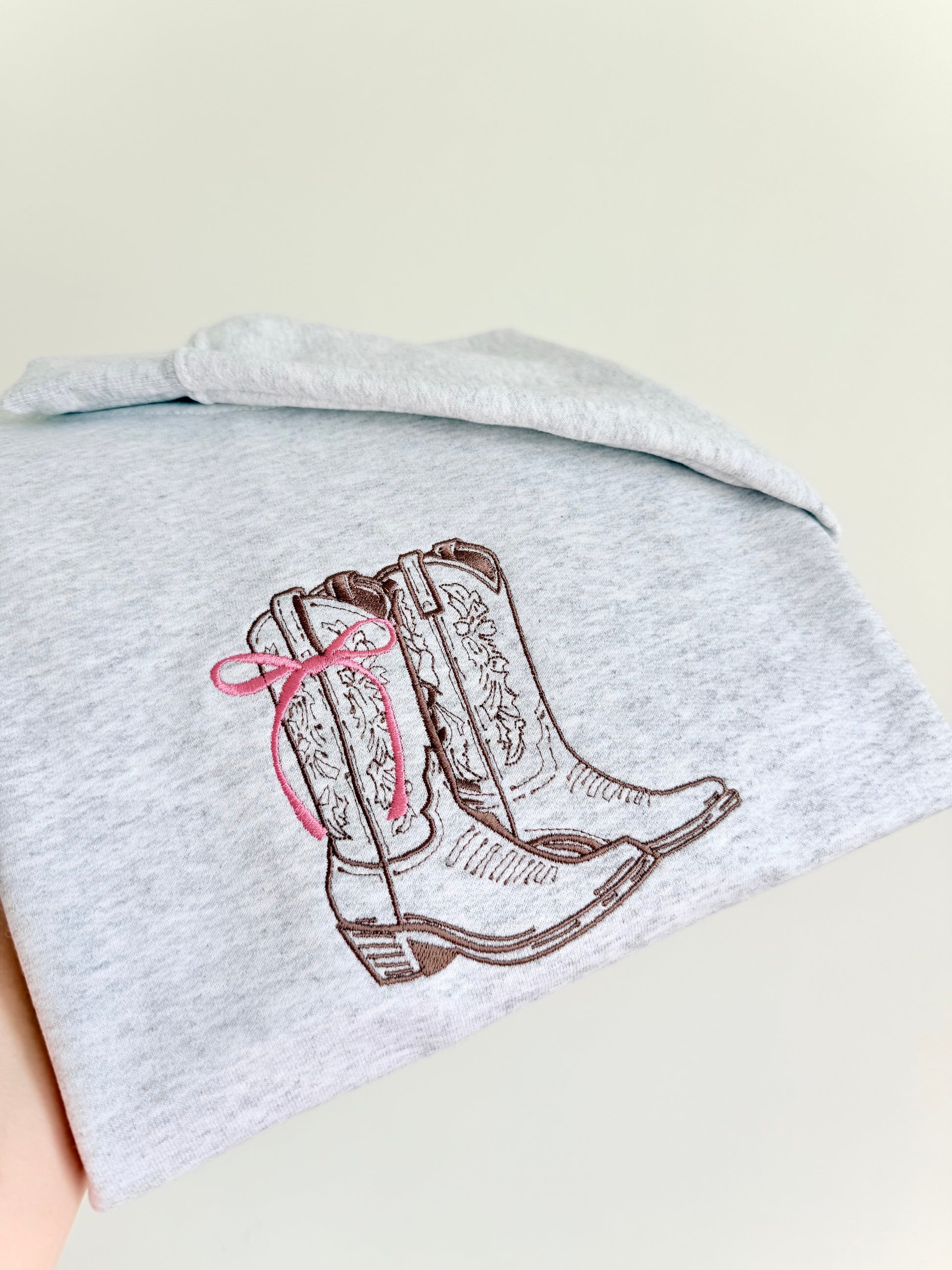 A folded light heather gray sweatshirt or hoodie with an embroidered design of cowboy boots in brown and a pink bow accent, held by a hand against a plain light background.