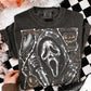 A folded black graphic T-shirt with a large white Ghostface print and Halloween-themed embellishments, held by a hand over a black-and-white checkered surface with fluffy white decor.