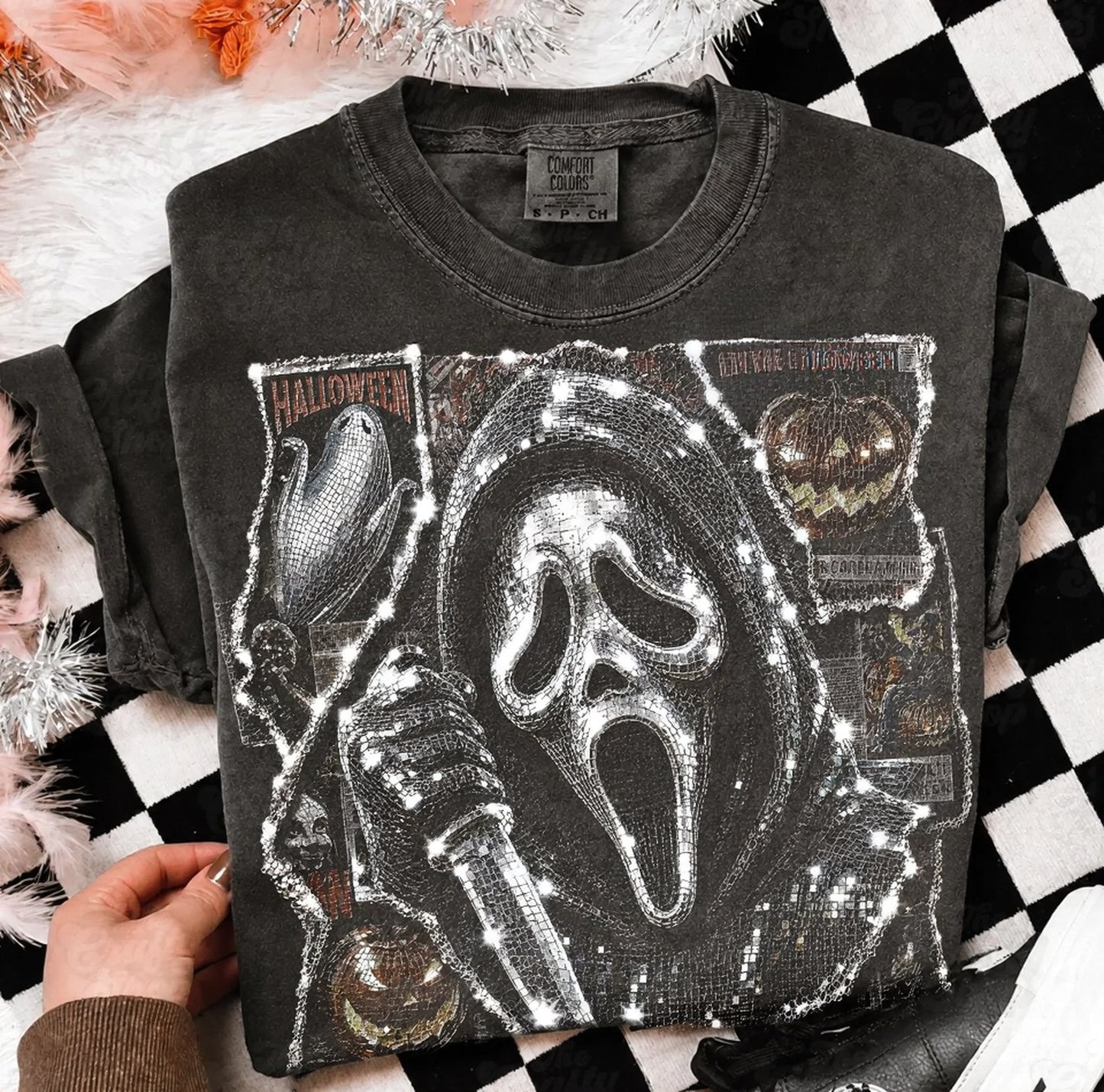 A folded black graphic T-shirt with a large white Ghostface print and Halloween-themed embellishments, held by a hand over a black-and-white checkered surface with fluffy white decor.