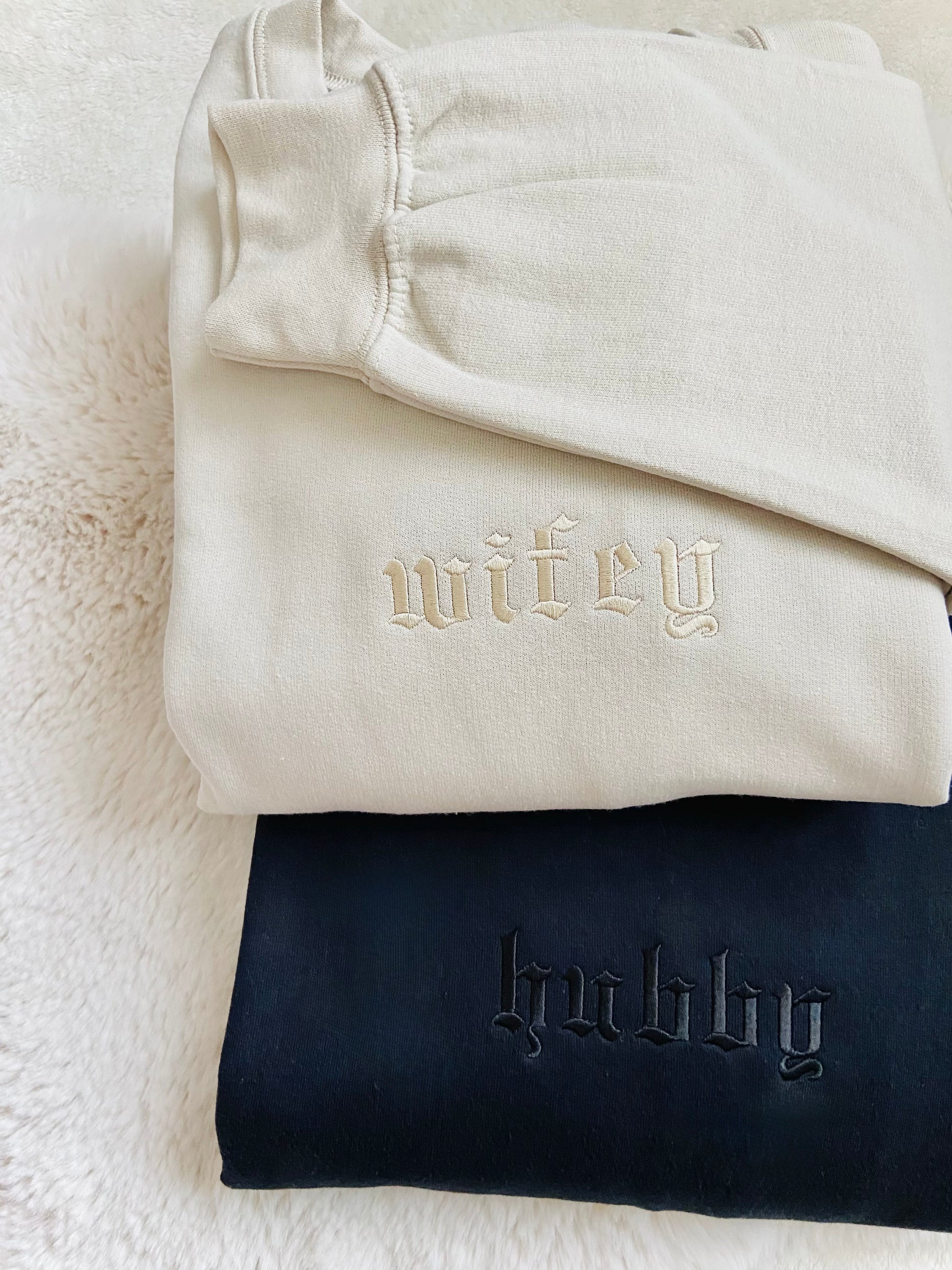 Folded beige sweatshirt with 'wifey' and navy sweatshirt with 'hubby' on a textured surface