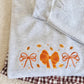 A folded light heather gray sweatshirt with orange embroidered bows and small leaf motifs, placed on top of a brown and white gingham fabric piece over a cream sherpa background.