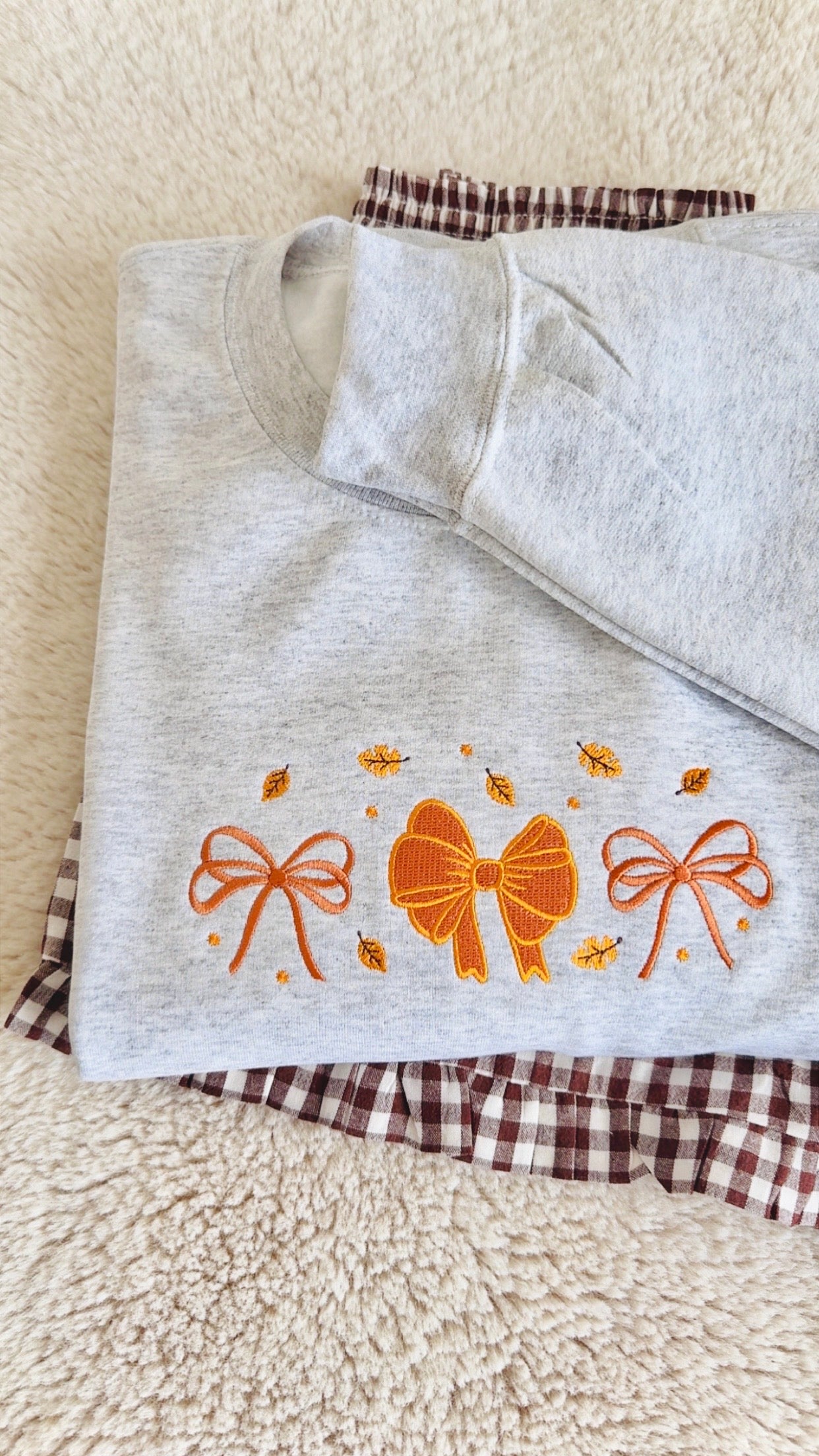 A folded light heather gray sweatshirt with orange embroidered bows and small leaf motifs, placed on top of a brown and white gingham fabric piece over a cream sherpa background.