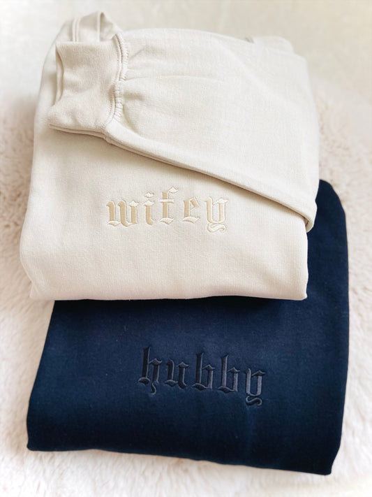 Folded white sweatshirt with 'wife' and navy sweatshirt with 'hubby' text on a textured surface.