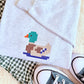 A folded light heather gray sweatshirt with a colorful pixelated MALLARD duck embroidery on the chest, styled on a cream faux fur surface with a green and white gingham shirt and black canvas sneakers.