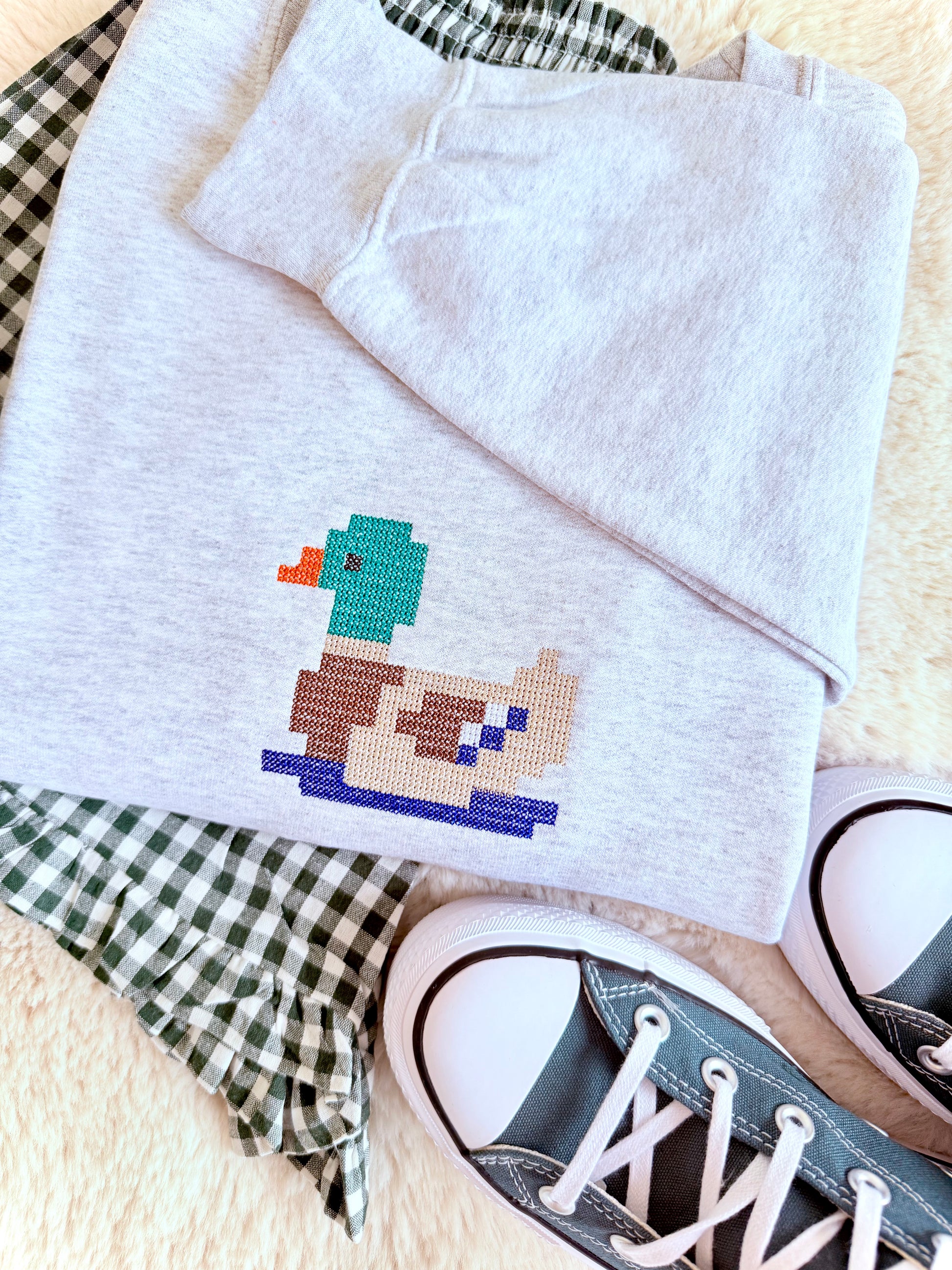 A folded light heather gray sweatshirt with a colorful pixelated MALLARD duck embroidery on the chest, styled on a cream faux fur surface with a green and white gingham shirt and black canvas sneakers.