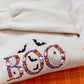 A folded cream-colored sweatshirt featuring a colorful embroidered 'BOO' design with bats, skulls, and pumpkins, layered over an orange plaid fabric.