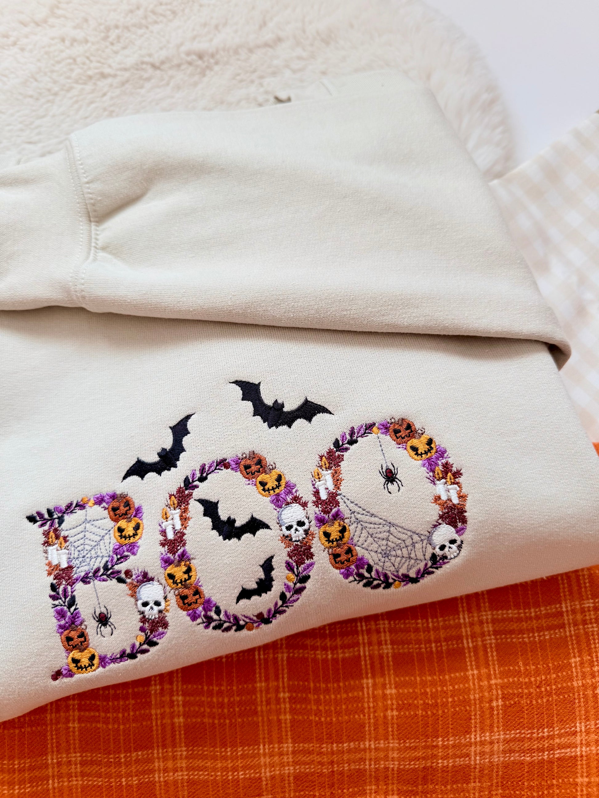 A folded cream-colored sweatshirt featuring a colorful embroidered 'BOO' design with bats, skulls, and pumpkins, layered over an orange plaid fabric.