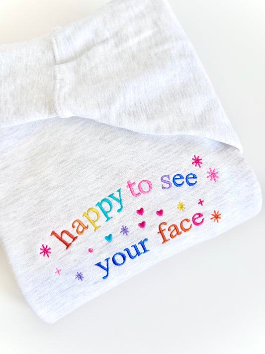 Grey sweater embroidered with colorful text 'happy to see your face' on a white background