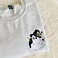 A folded light heather gray t-shirt with a small embroidered cartoon ghost wearing a black witch hat on the chest, displayed on a soft cream-colored faux fur surface.