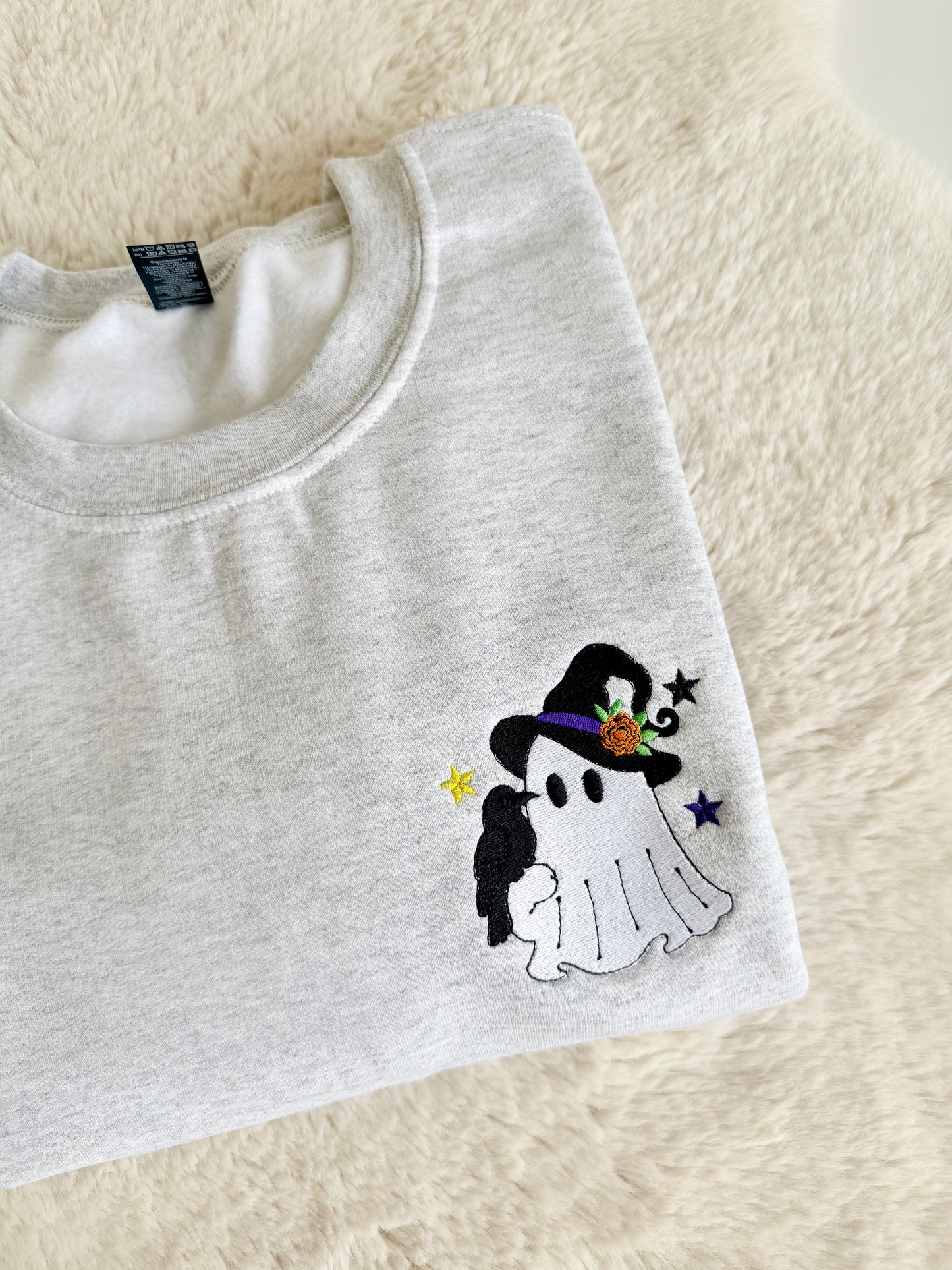 A folded light heather gray t-shirt with a small embroidered cartoon ghost wearing a black witch hat on the chest, displayed on a soft cream-colored faux fur surface.
