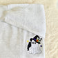 A folded light gray heather sweatshirt on a soft cream faux-fur surface, featuring a small embroidered Halloween ghost design with a black witch hat, purple band, orange flower, black bird, and small stars.