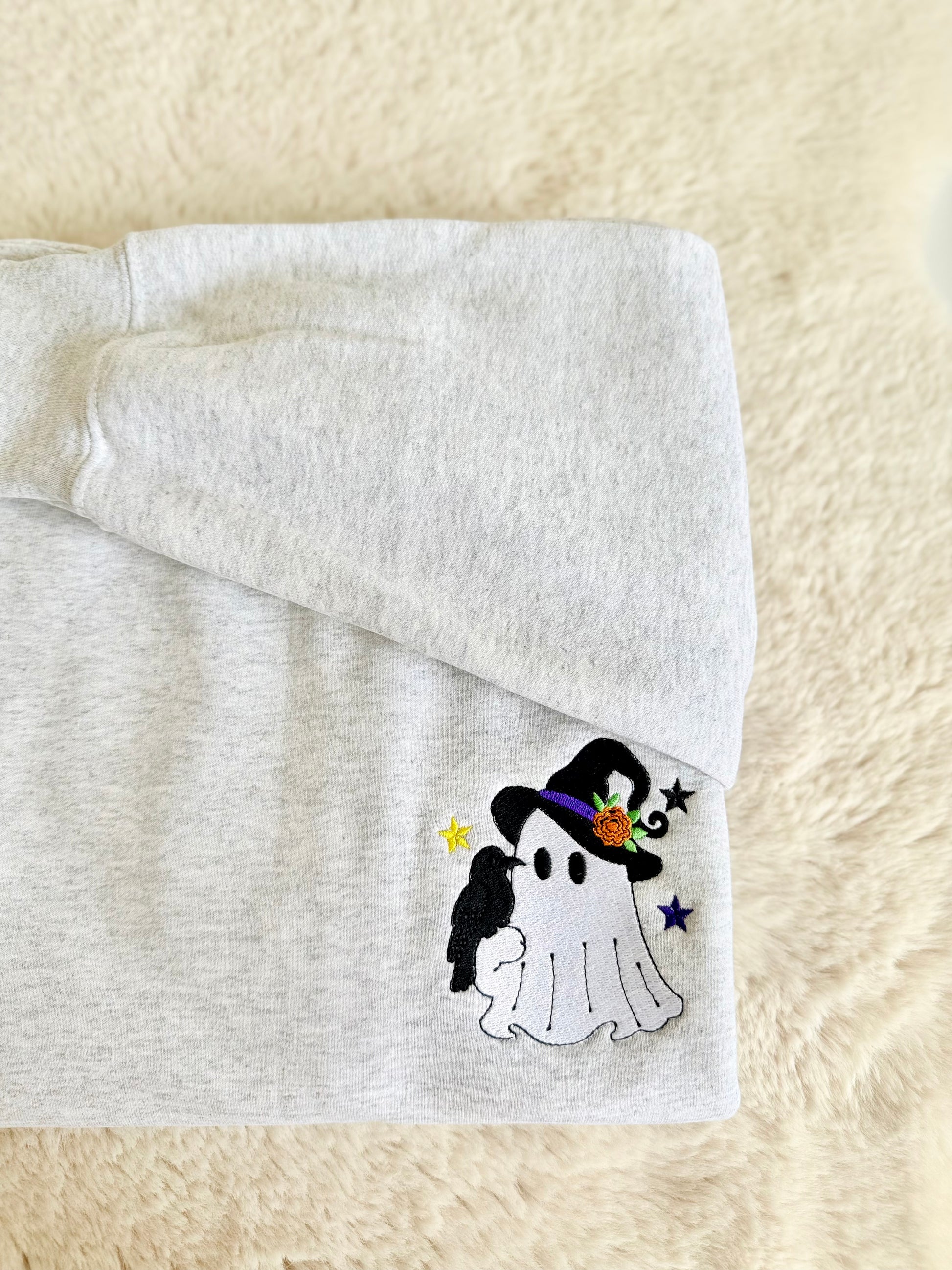 A folded light gray heather sweatshirt on a soft cream faux-fur surface, featuring a small embroidered Halloween ghost design with a black witch hat, purple band, orange flower, black bird, and small stars.