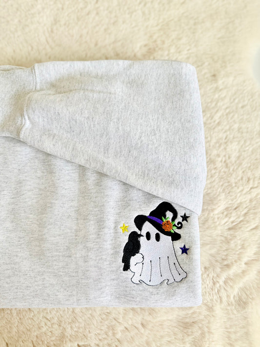 A folded light gray heather sweatshirt on a soft cream faux-fur surface, featuring a small embroidered Halloween ghost design with a black witch hat, purple band, orange flower, black bird, and small stars.