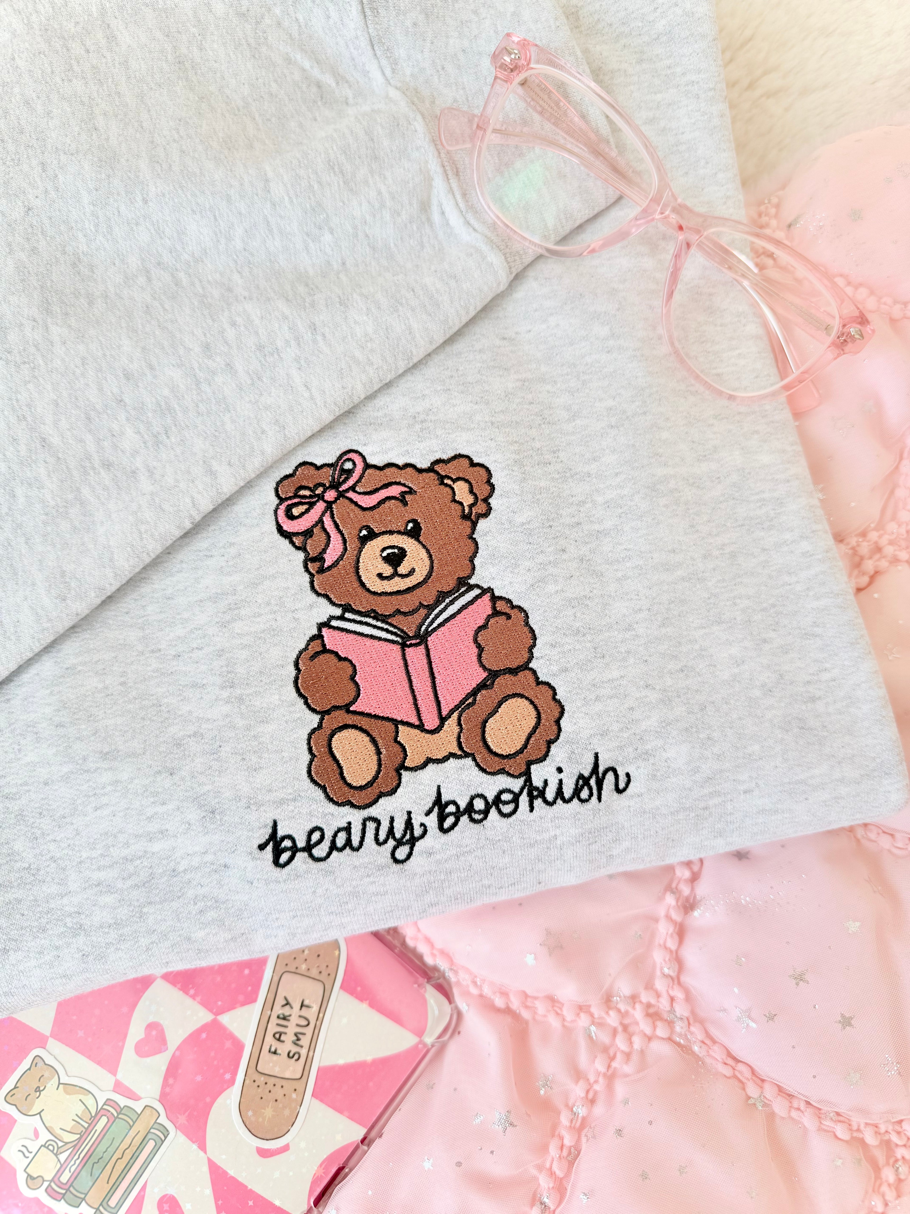 Gray sweatshirt with a teddy bear reading a book design and 'bearly bookish' text, surrounded by pink and white items.