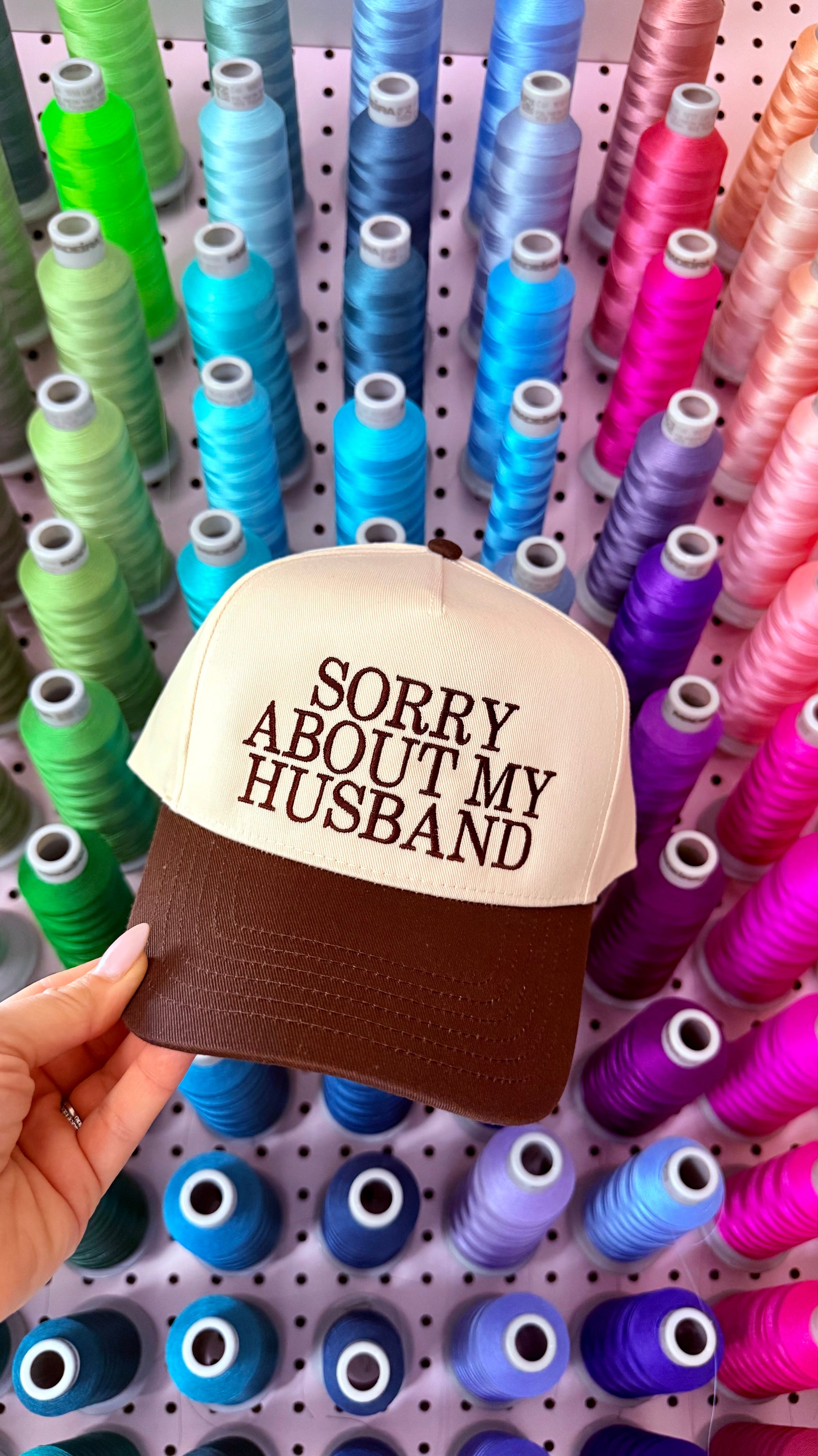 Cap with 'Sorry About My Husband' text held in front of spools of thread.