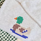 A light heather gray sweatshirt featuring a colorful pixelated duck design on the front, styled on a green and white gingham blanket.