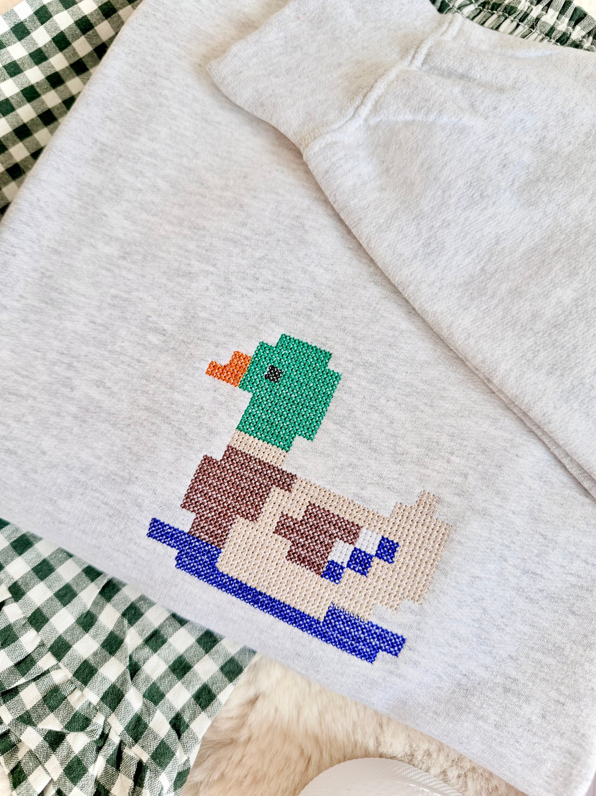 A light heather gray sweatshirt featuring a colorful pixelated duck design on the front, styled on a green and white gingham blanket.