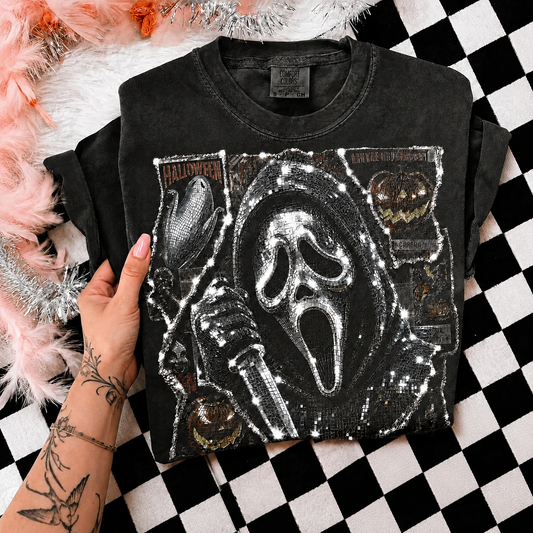 A black sequined t-shirt featuring a Ghostface graphic, held by a tattooed hand over a black and white checkered surface with pink and white faux fur props.