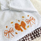 A folded light heather gray garment, likely a hoodie or sweatshirt, featuring embroidered orange bows and small yellow leaves on the front. A brown and white gingham fabric is partially visible underneath.