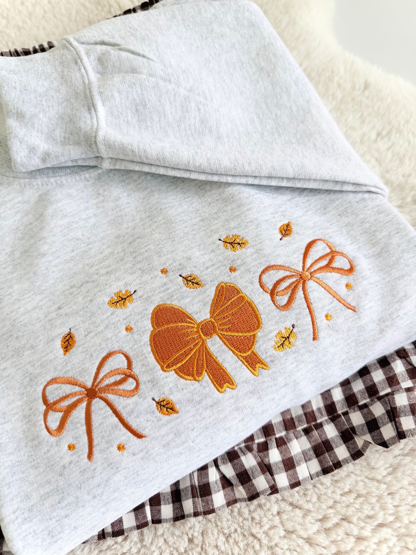 A folded light heather gray garment, likely a hoodie or sweatshirt, featuring embroidered orange bows and small yellow leaves on the front. A brown and white gingham fabric is partially visible underneath.