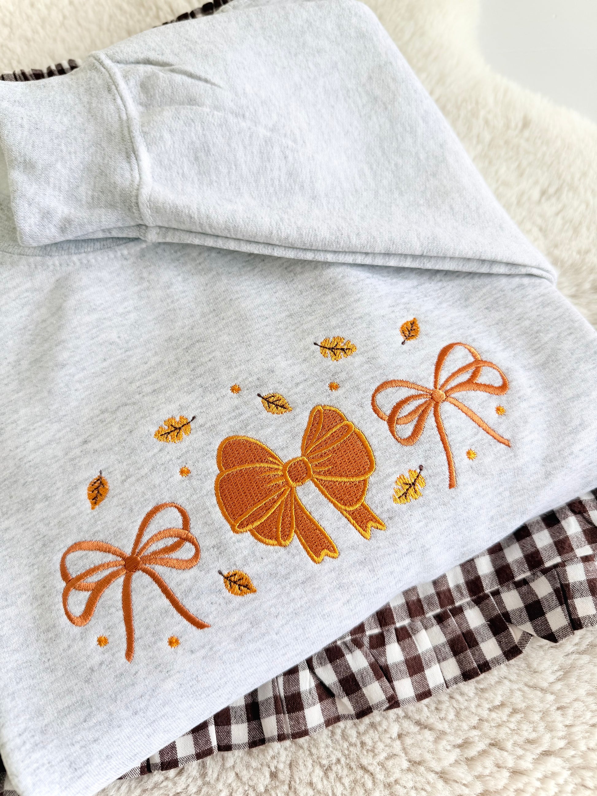 A folded light heather gray garment, likely a hoodie or sweatshirt, featuring embroidered orange bows and small yellow leaves on the front. A brown and white gingham fabric is partially visible underneath.