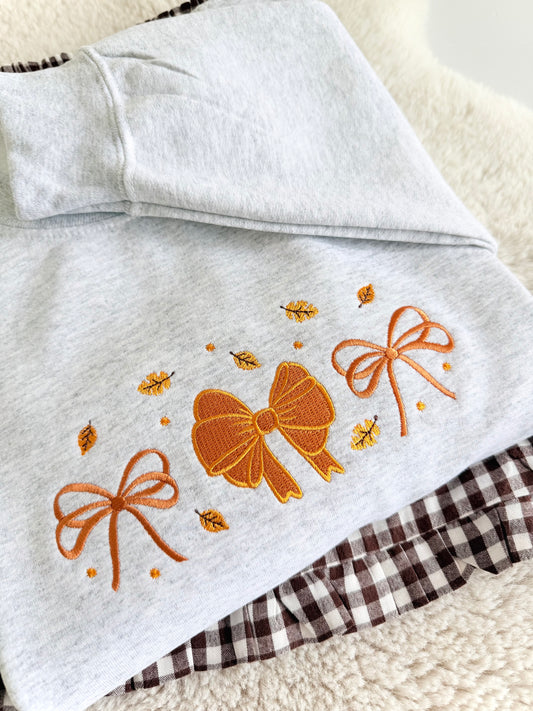A folded light heather gray garment, likely a hoodie or sweatshirt, featuring embroidered orange bows and small yellow leaves on the front. A brown and white gingham fabric is partially visible underneath.
