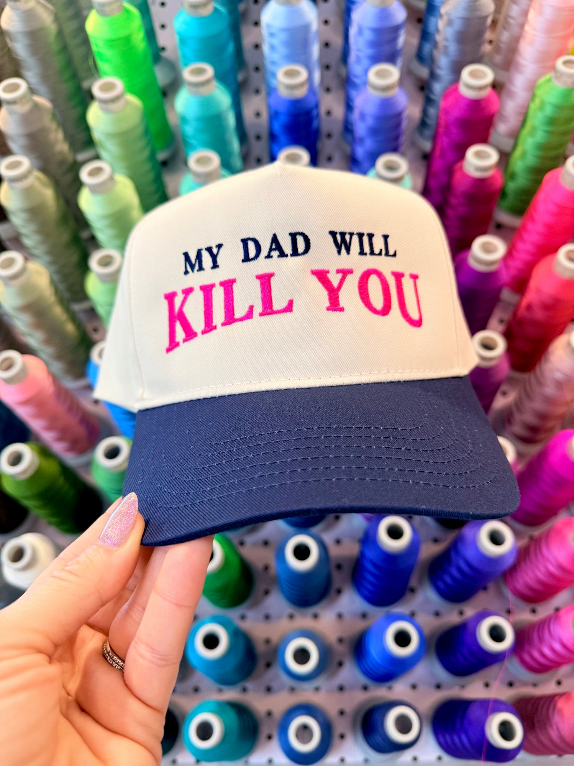 Cap with 'My Dad Will Kill You' text held in front of spools of colorful thread.