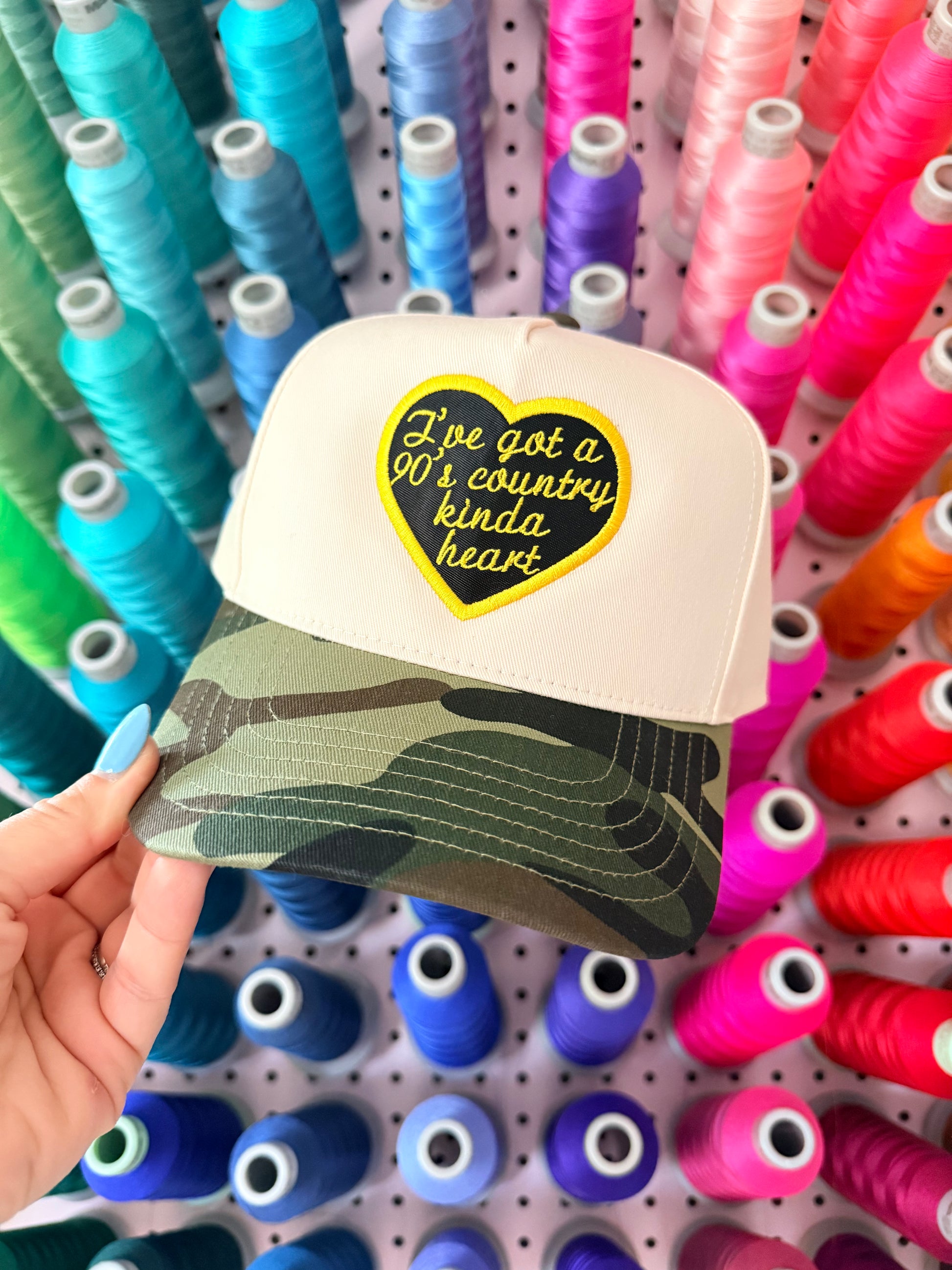 Cap with heart-shaped patch held in front of spools of colourful thread. 90's inspired millennial embroidered snapback hat.