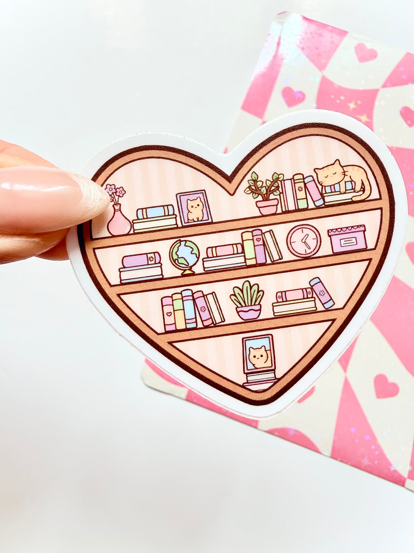 Heart-shaped sticker with a bookshelf design held by a hand on a pink heart-patterned background.