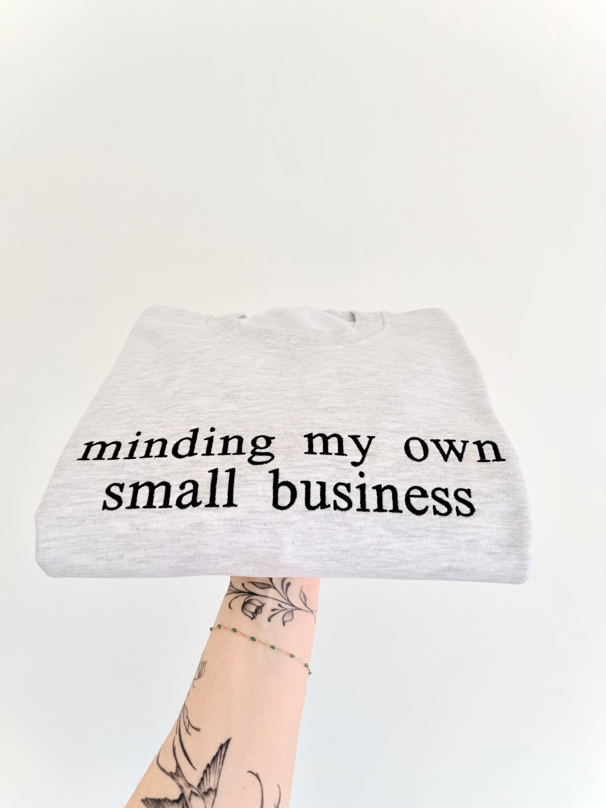light ash grey crewneck with 'minding my own small business' text embroidered on it, displayed on a white background.