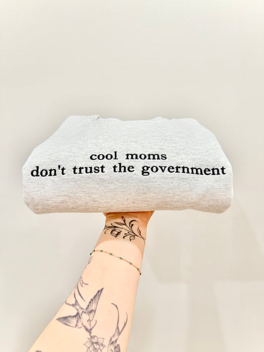 Sweater with 'cool moms don't trust the government' text embroidered held by a person with tattoos on a light background