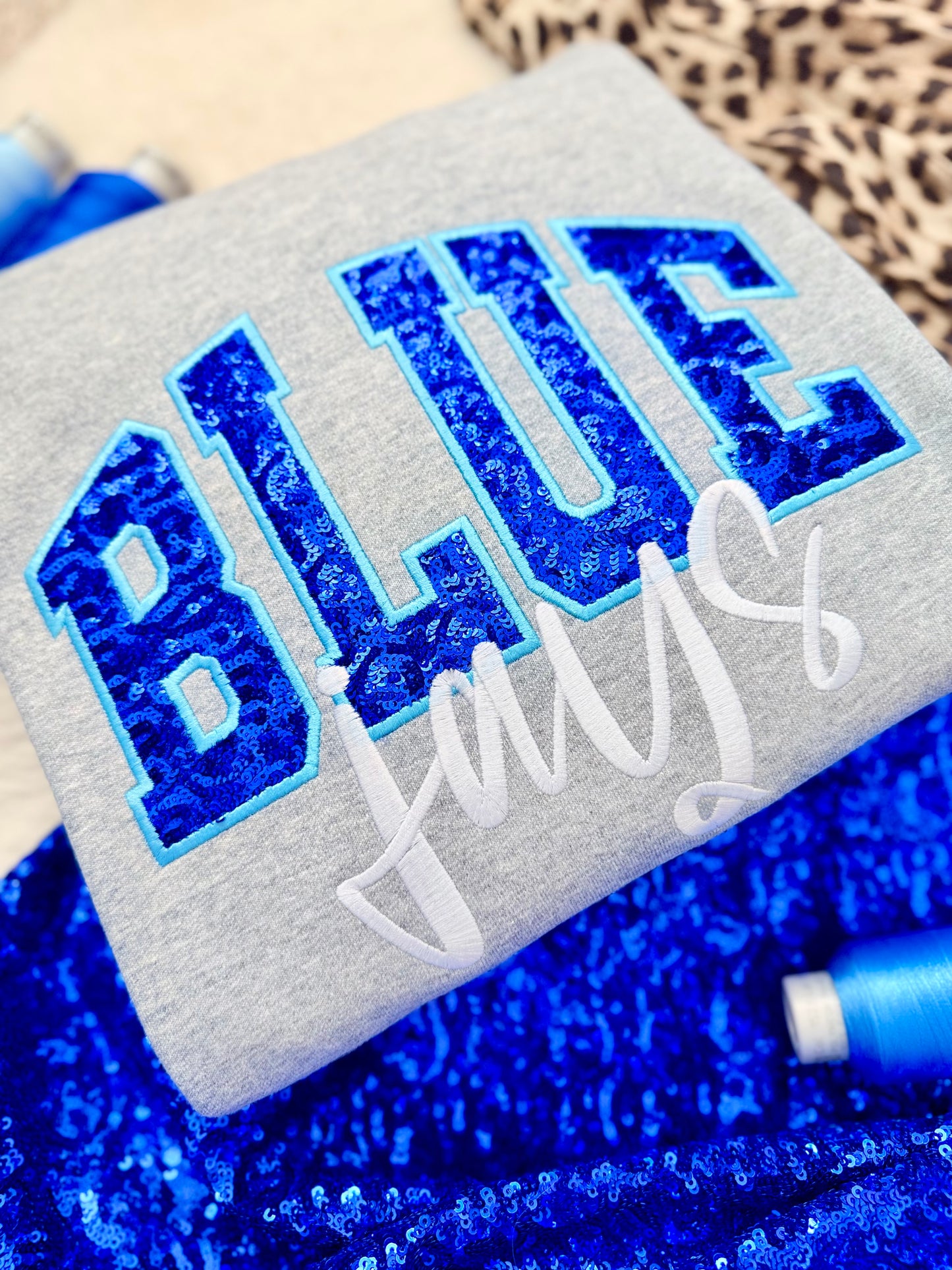 Gray sweater with blue glittery text and white cursive text on a blue textured background. Blue Jays fanwear.