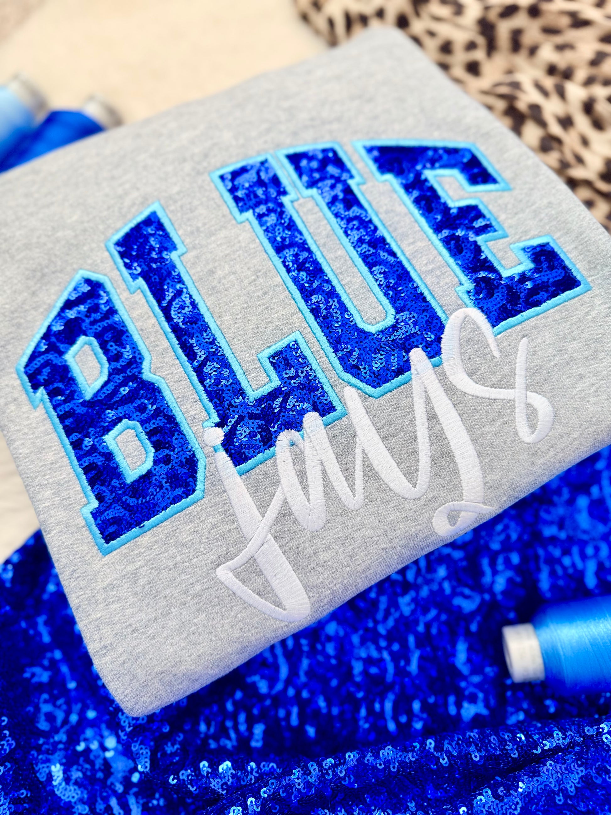 Gray sweater with blue glittery text and white cursive text on a blue textured background. Blue Jays fanwear.