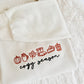 An off-white hooded sweatshirt with embroidered autumn-themed icons and the words 'cogy season' in red and black, laid flat on a white textured blanket.