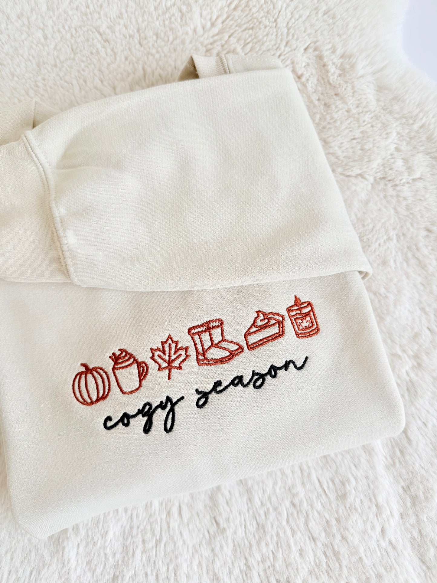 An off-white hooded sweatshirt with embroidered autumn-themed icons and the words 'cogy season' in red and black, laid flat on a white textured blanket.