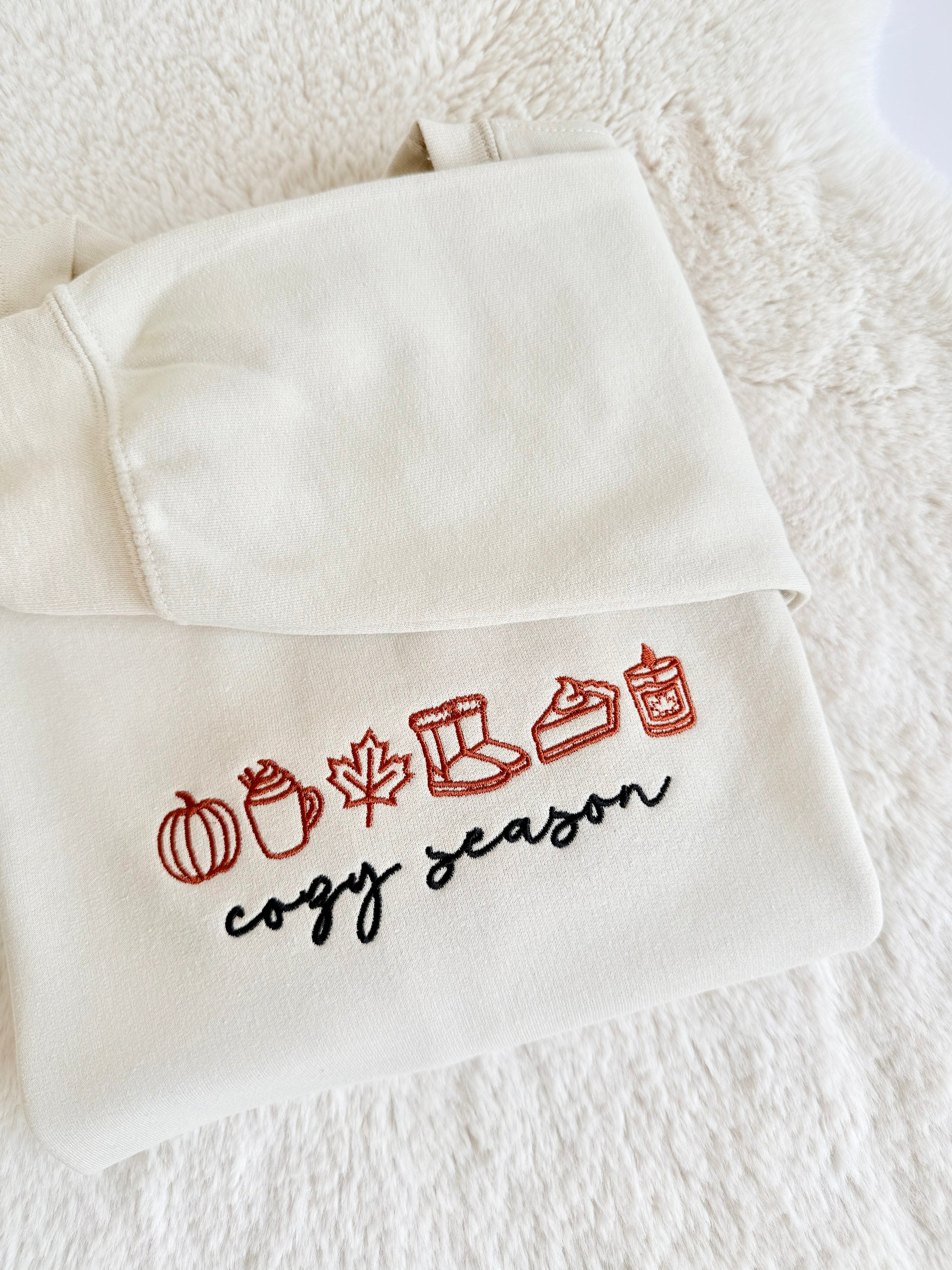 An off-white hooded sweatshirt with embroidered autumn-themed icons and the words 'cogy season' in red and black, laid flat on a white textured blanket.