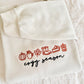 A folded cream-colored sweatshirt or hoodie laid flat on a soft white textured blanket. The front has embroidered autumn-themed icons in red and black text reading 'crazy season'.