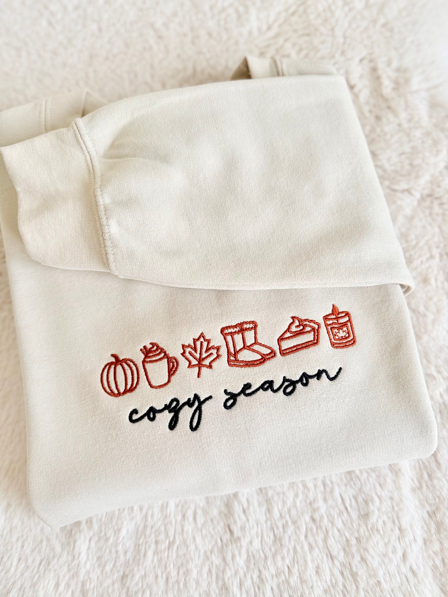 A folded cream-colored sweatshirt or hoodie laid flat on a soft white textured blanket. The front has embroidered autumn-themed icons in red and black text reading 'crazy season'.