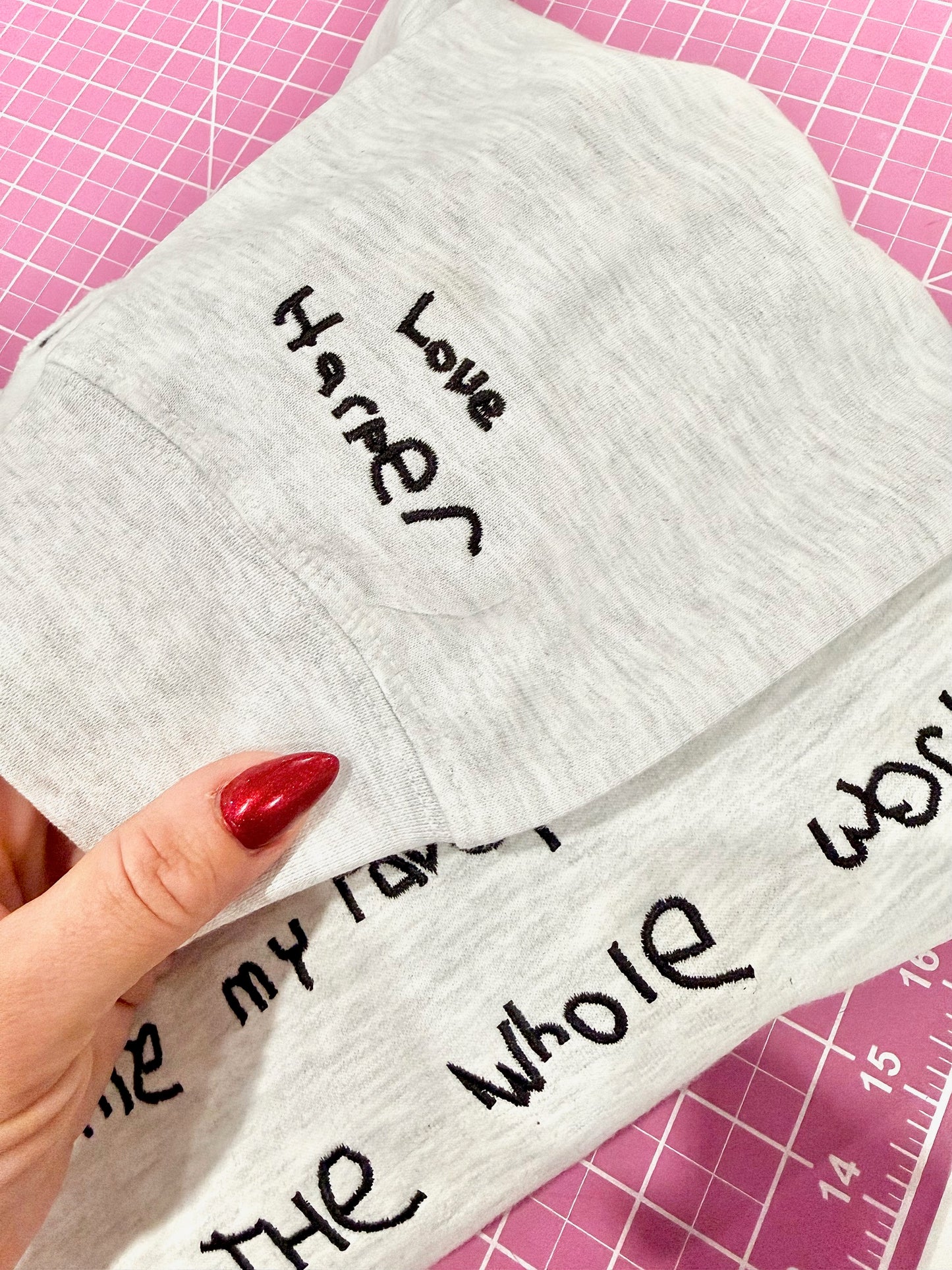 Gray fabric with embroidered text held by a hand on a pink cutting mat.