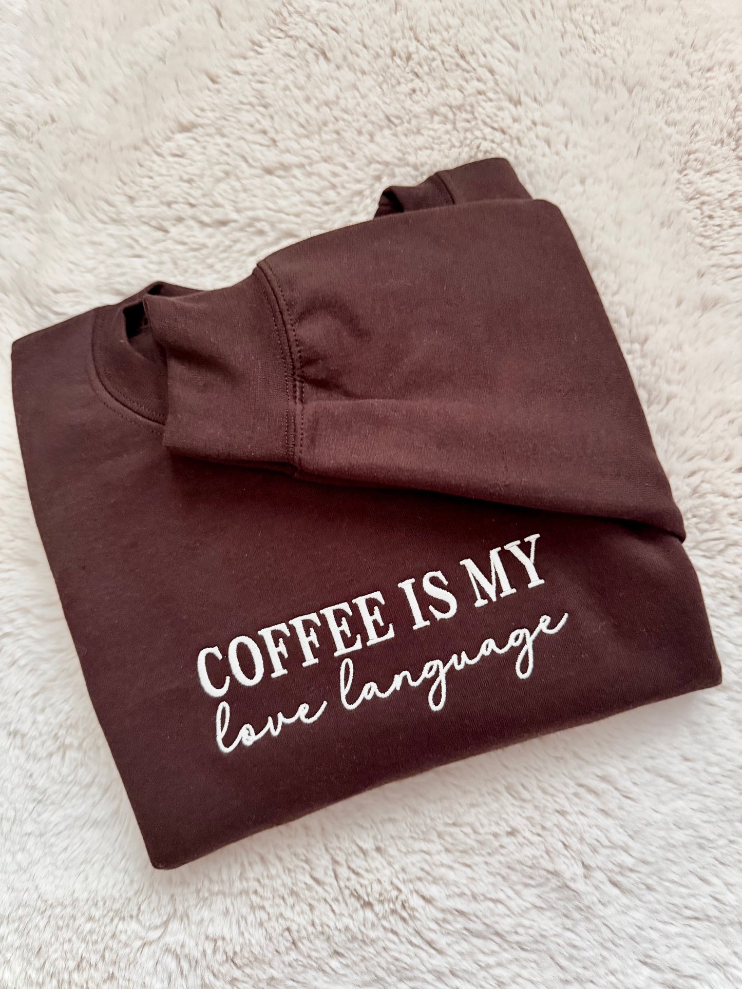 Folded brown sweatshirt with 'Coffee is my love language' text on a textured white surface