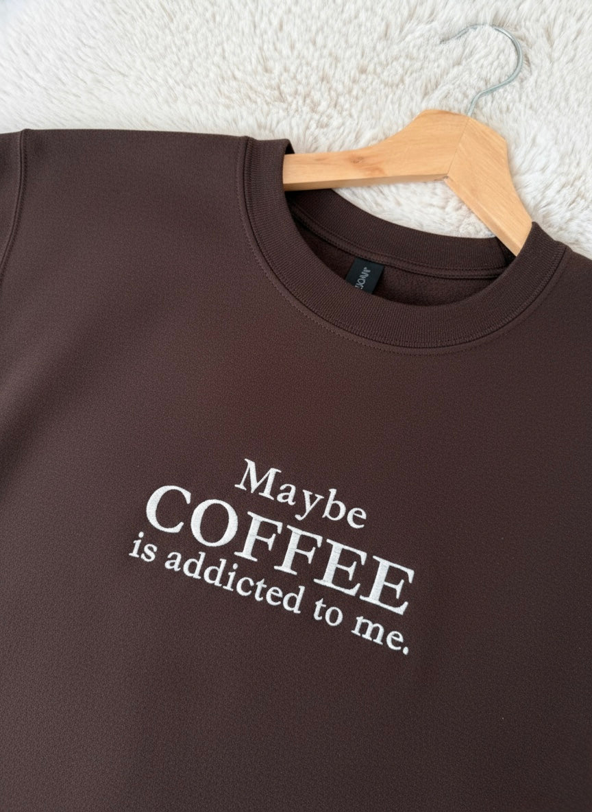 Brown sweatshirt with 'Maybe COFFEE is addicted to me.' text on a hanger against a textured white background