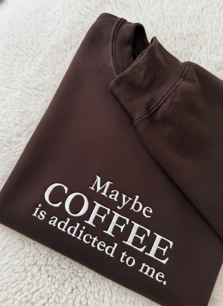 Brown sweatshirt with text 'Maybe COFFEE is addicted to me.' on a textured surface