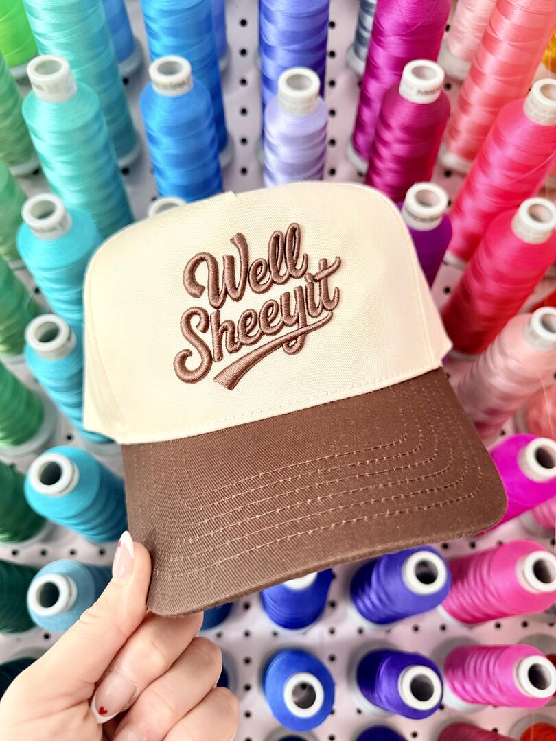 Cap with 'Well Sheeyit' text held in front of spools of colorful thread.