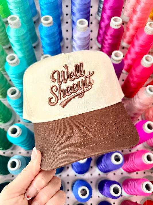 Cap with 'Well Sheeyit' text held in front of spools of colorful thread.